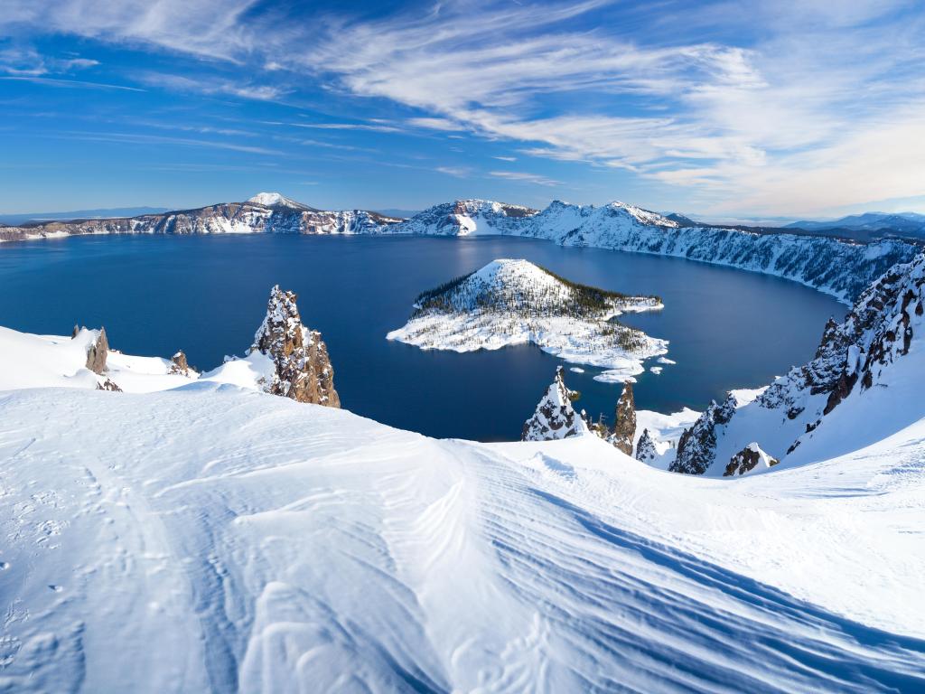 View of blue lake in volcano crater with snow-covered rim and mountain sides and blue sky