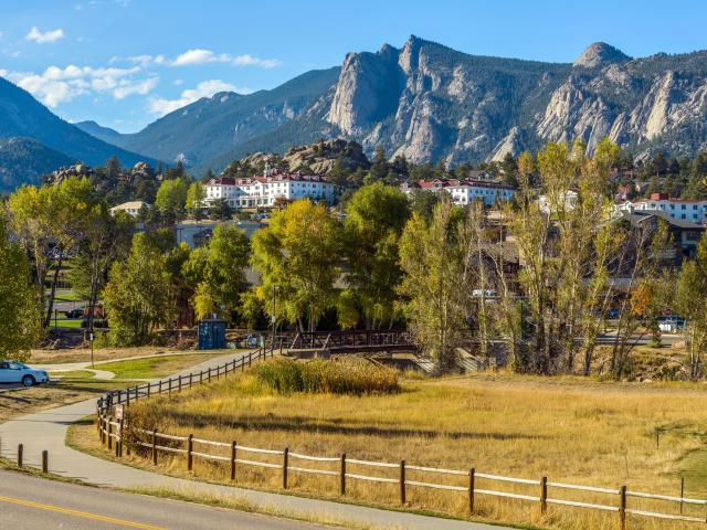 A road winds towards Downtown Estes Park, with The Stanley Hotel and Rocky Mountains in background on a sunny fall afternoon