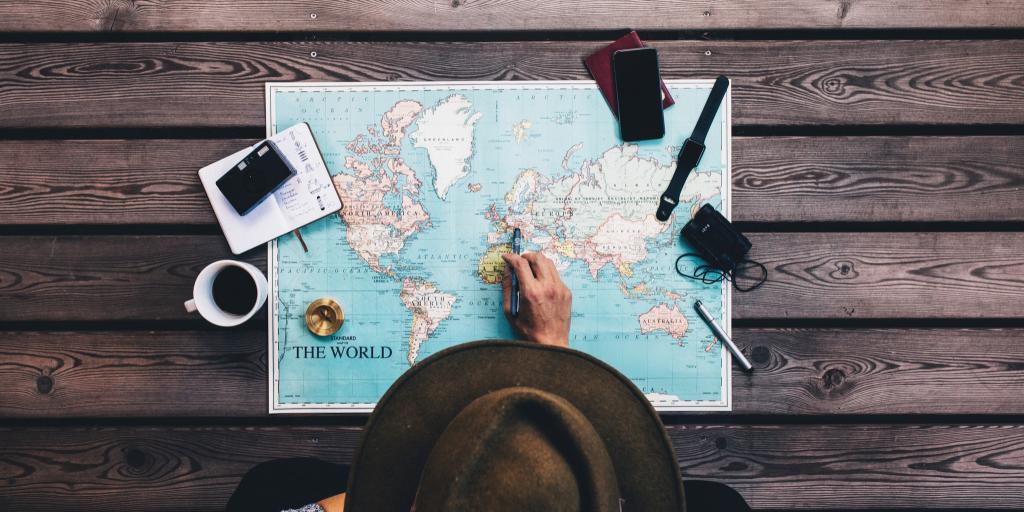 Man in a hat sits at a table looking at a map with binoculars, compass and other travel accessories