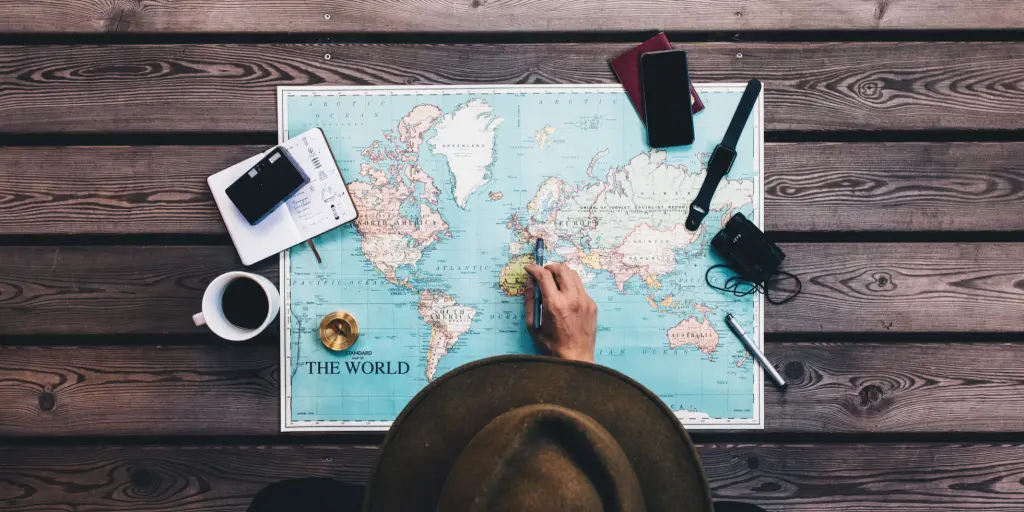 Man in a hat sits at a table looking at a map with binoculars, compass and other travel accessories