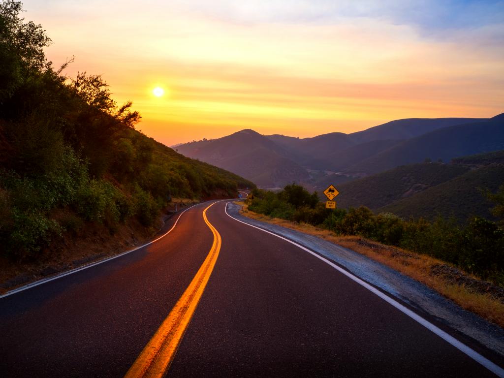 A scenic view of the mountains at Old Priest Grade Road with green grasses and trees at the side of the road with a beautiful orange-yellow sunset