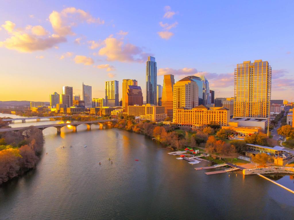 The Austin, Texas skyline cityscape under a sunset sky is beautifully captured in an aerial landscape, with the city's landmark buildings casting reflections on the serene Ladybird Lake - a popular tourist destination.