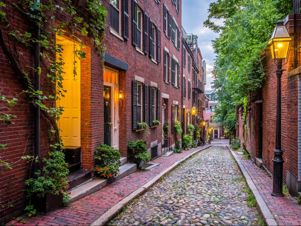 Boston, Massachusetts, USA with a view of Acorn Street, a pretty road with cobbled stones, street lights and pretty plants lining the street.