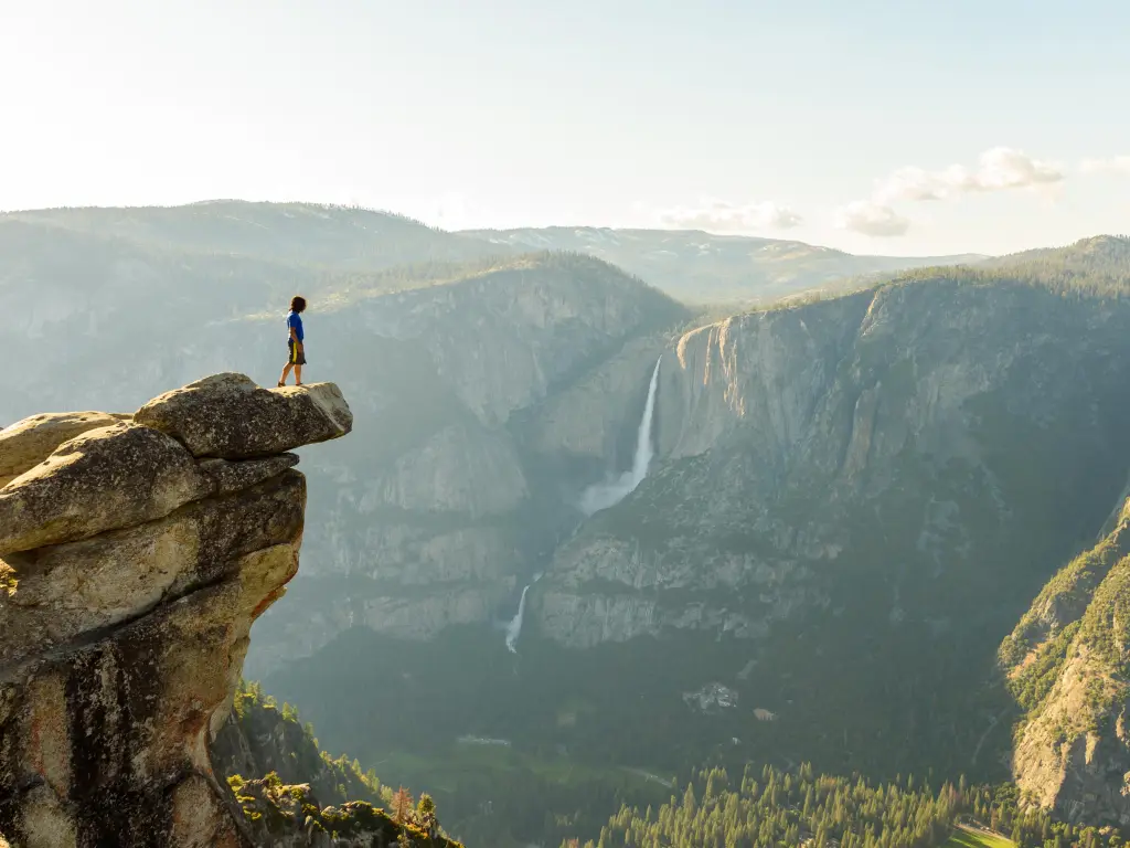 Hiker standing on a rock over the cliff on a sunny day with Yosemite Falls in the background