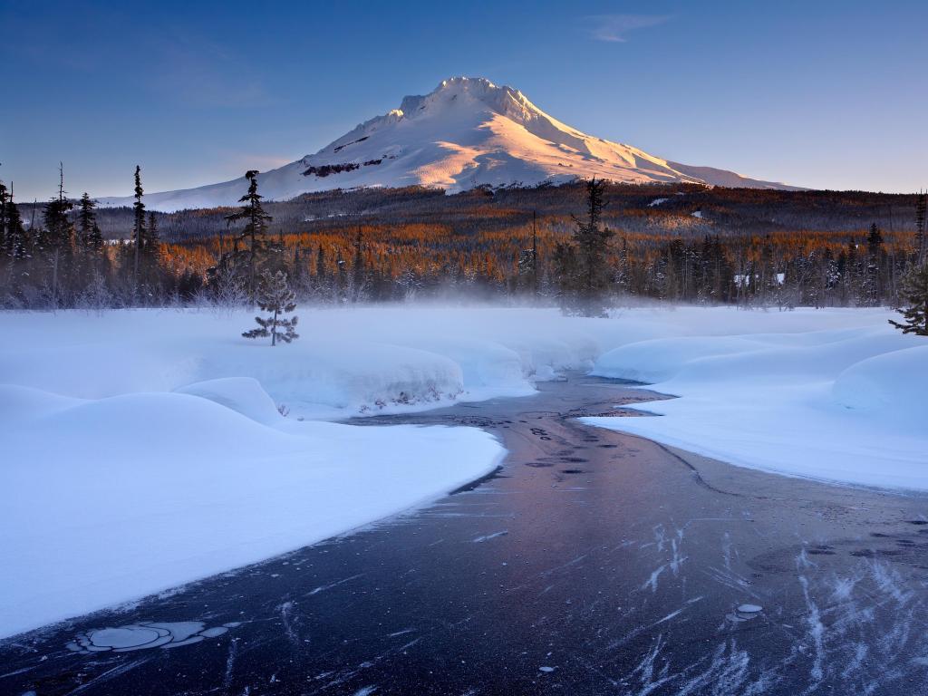 Snow covered mountain rising up in the distance behind pine trees and a frozen river with snowy banks