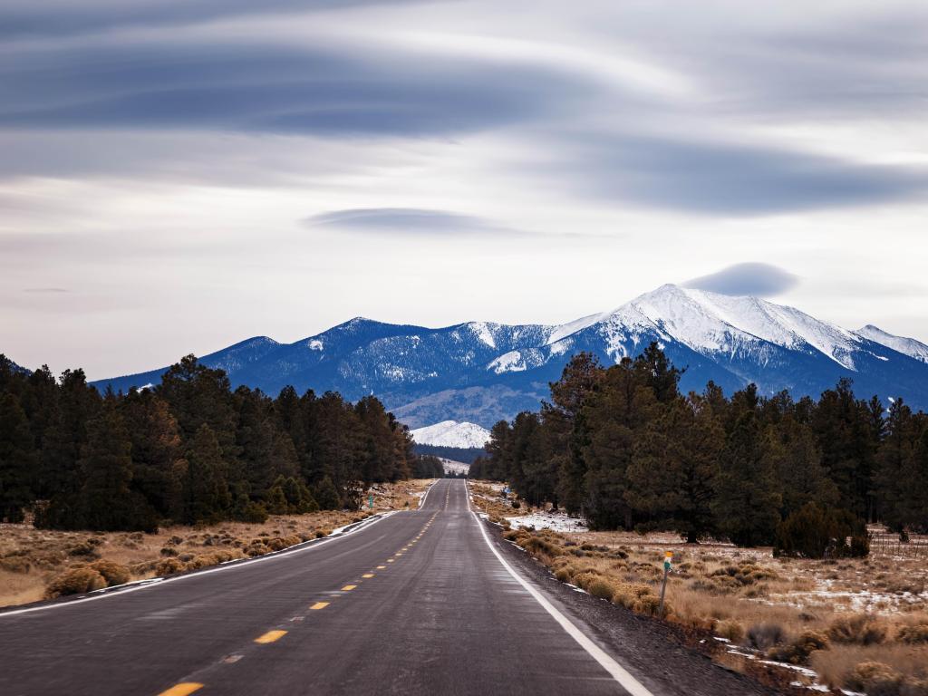 Flagstaff, Arizona with clouds over the snow covered San Francisco Peak in the distance and a road leading towards the distance.