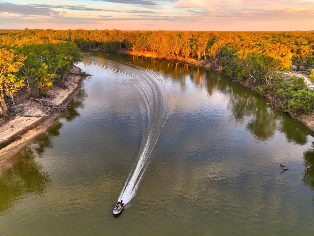 Sun setting over the trees, with a river in the middle of the image, speedboat sailing through