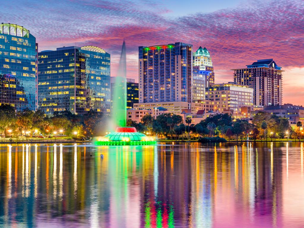 Fountain jet lit green shoots up from still lake water with cityscape and colorful cotton wool sunset sky