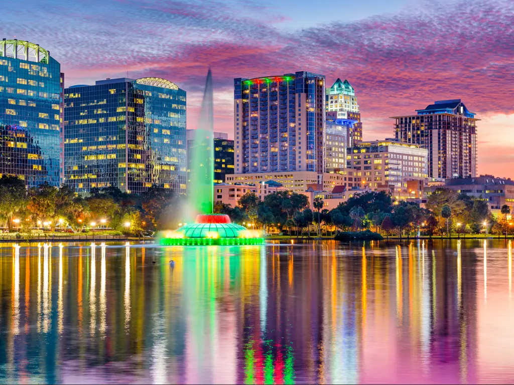 Fountain jet lit green shoots up from still lake water with cityscape and colorful cotton wool sunset sky