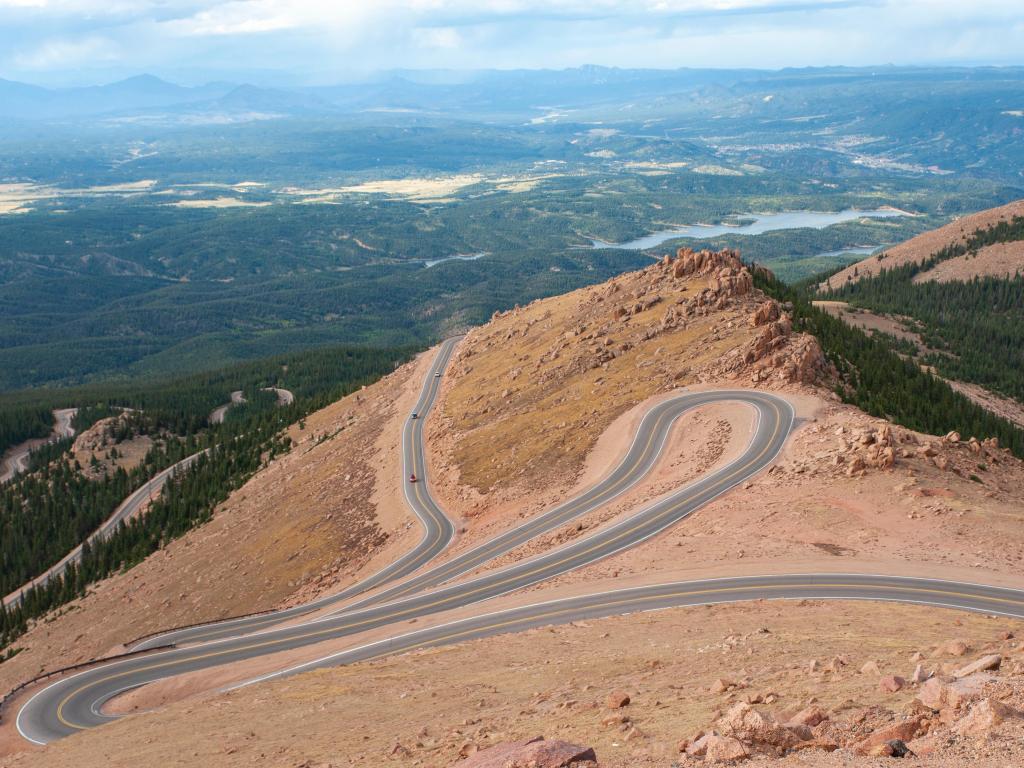 A winding road leading down from Pikes Peak in Colorado Springs with forests and hills in the background on a slightly overcast day.