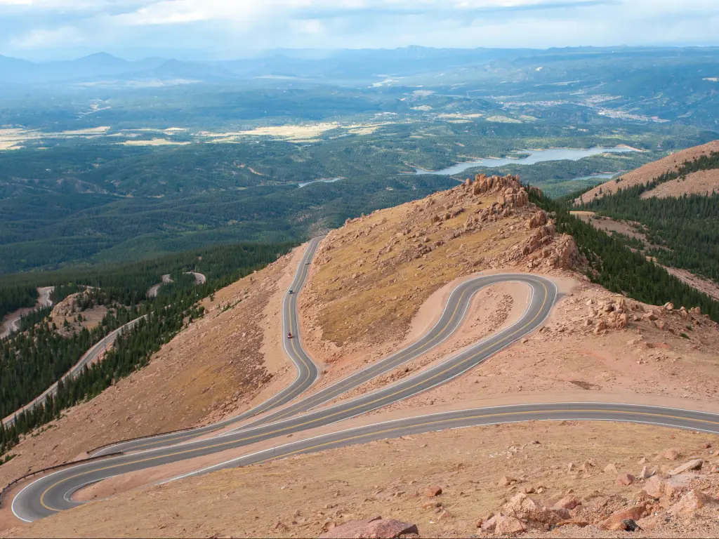 A winding road leading down from Pikes Peak in Colorado Springs with forests and hills in the background on a slightly overcast day.