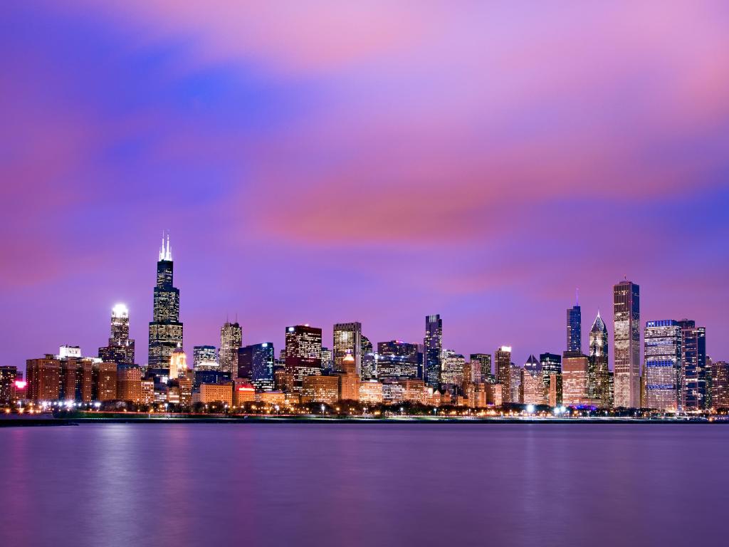 High rise buildings lit up at night with dark blue and pink sky and grey blue lake