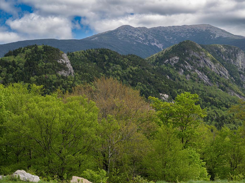 White Mountain National Forest in New Hampshire, USA with green trees in the foreground and the mountains in the distance on a sunny and cloudy day. 