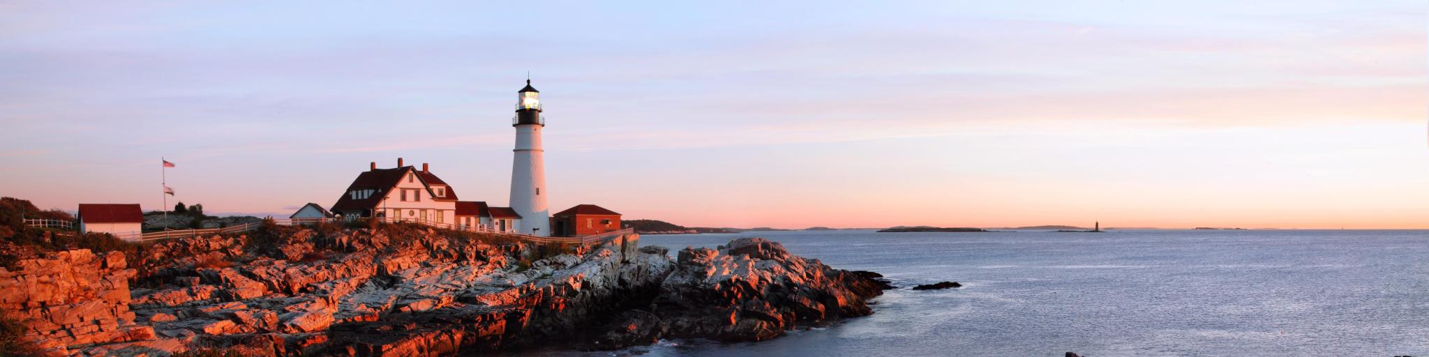 Panorama of Portland Head Lighthouse at dawn's first light, casting a warm glow on the rocks
