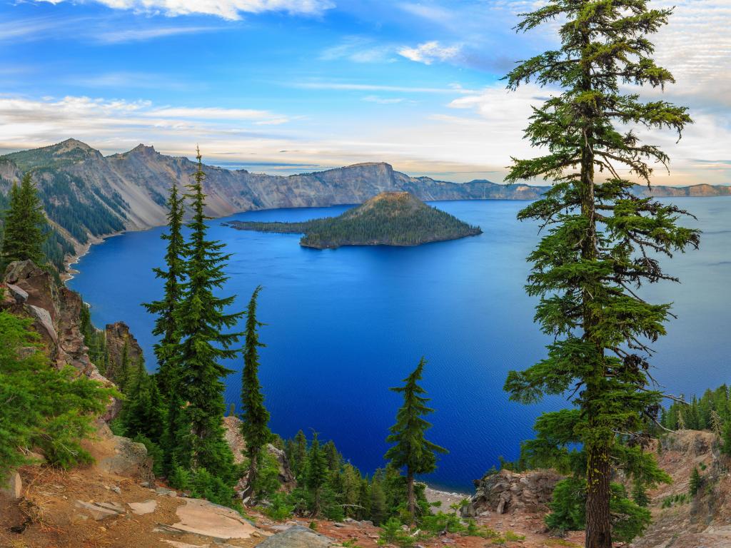 Crater Lake National Park, Oregon, USA with trees and rocks in the distance, the blue lake and mountains in the distance against a blue sky.