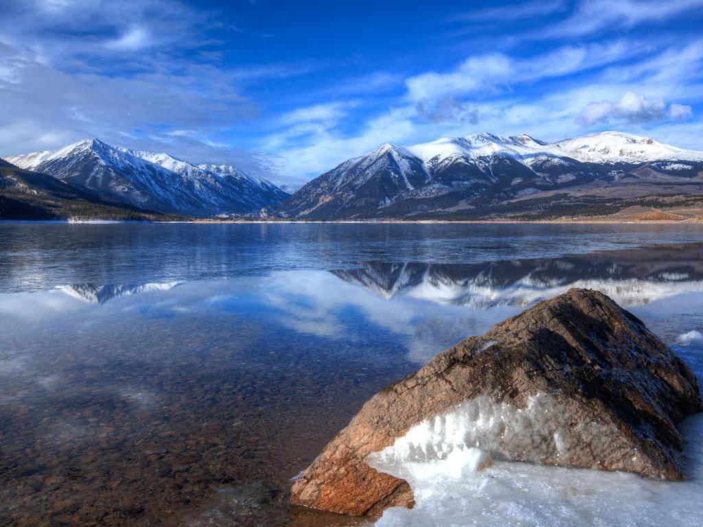 Twin Lakes, Colorado, USA with snow and rocks in the foreground, a calm lake and snow-capped mountains in the distance on a sunny clear day.