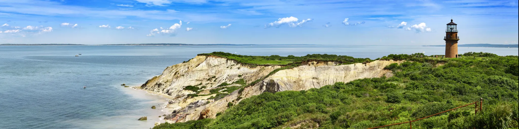 A panoramic view of the Coastal Lighthouse on a green grassy cliff with a mirroring sky and sea of blue with fluffy cotton-like clouds in Aquinnah, Martha's Vineyard
