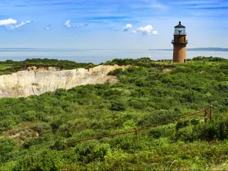 A panoramic view of the Coastal Lighthouse on a green grassy cliff with a mirroring sky and sea of blue with fluffy cotton-like clouds in Aquinnah, Martha's Vineyard