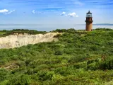 A panoramic view of the Coastal Lighthouse on a green grassy cliff with a mirroring sky and sea of blue with fluffy cotton-like clouds in Aquinnah, Martha's Vineyard