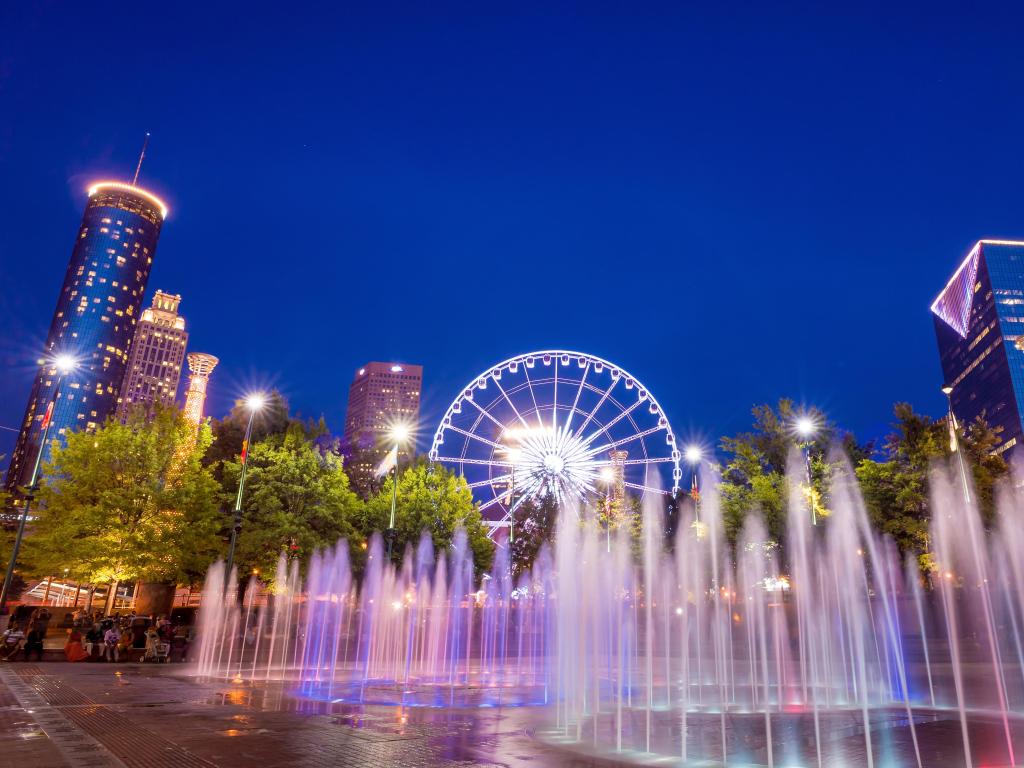 Fountains lit up pink and blue with ferris wheel and high rise buildings in the background against night sky