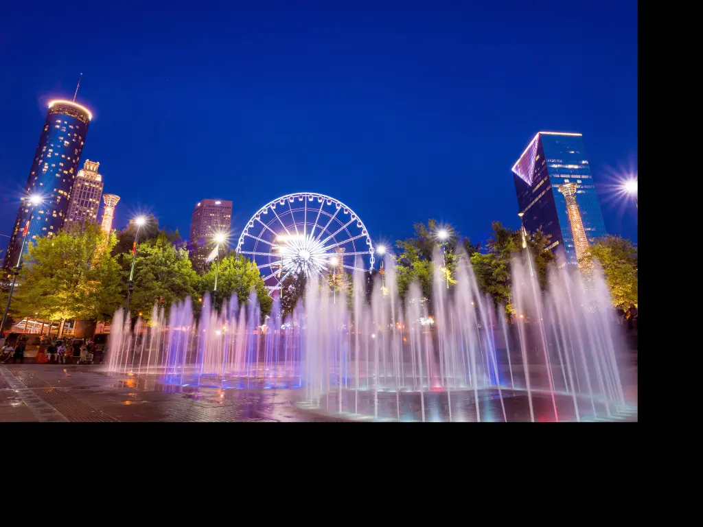 Fountains lit up pink and blue with ferris wheel and high rise buildings in the background against night sky