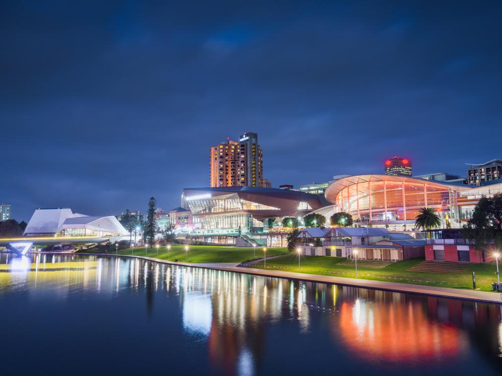 Adelaide, Australia at night with water in the foreground reflecting the buildings which are illuminated with lights and street lamps.