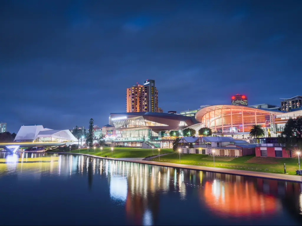 Adelaide, Australia at night with water in the foreground reflecting the buildings which are illuminated with lights and street lamps.