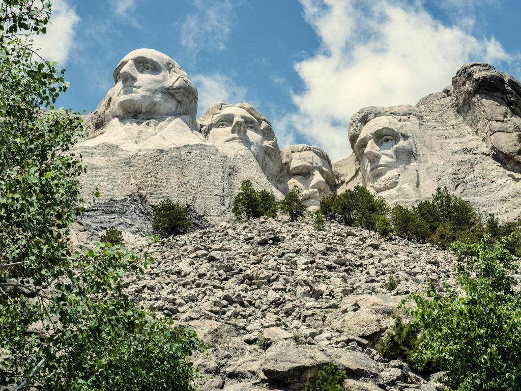 Mount Rushmore, South Dakota, USA with the famous presidents carved into Black Hills, trees in the foreground and a cloudy but sunny sky above.