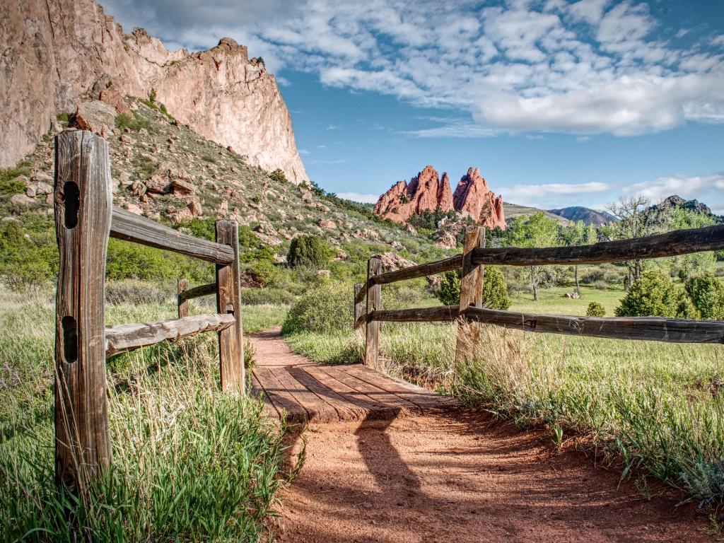 A hiking trail crosses a bridge in a meadow in Garden Of The Gods, Colorado with red sandstone rock formations in the background.