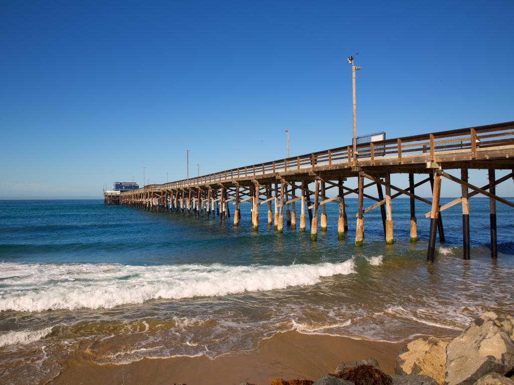 An image of a pier at New Port Beach on the sea and some rocks on the lower right side during a bright sunny day.