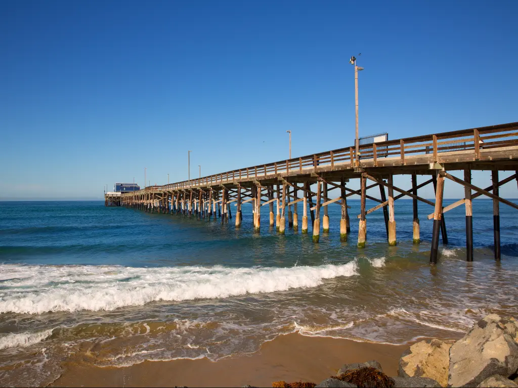 An image of a pier at New Port Beach on the sea and some rocks on the lower right side during a bright sunny day.