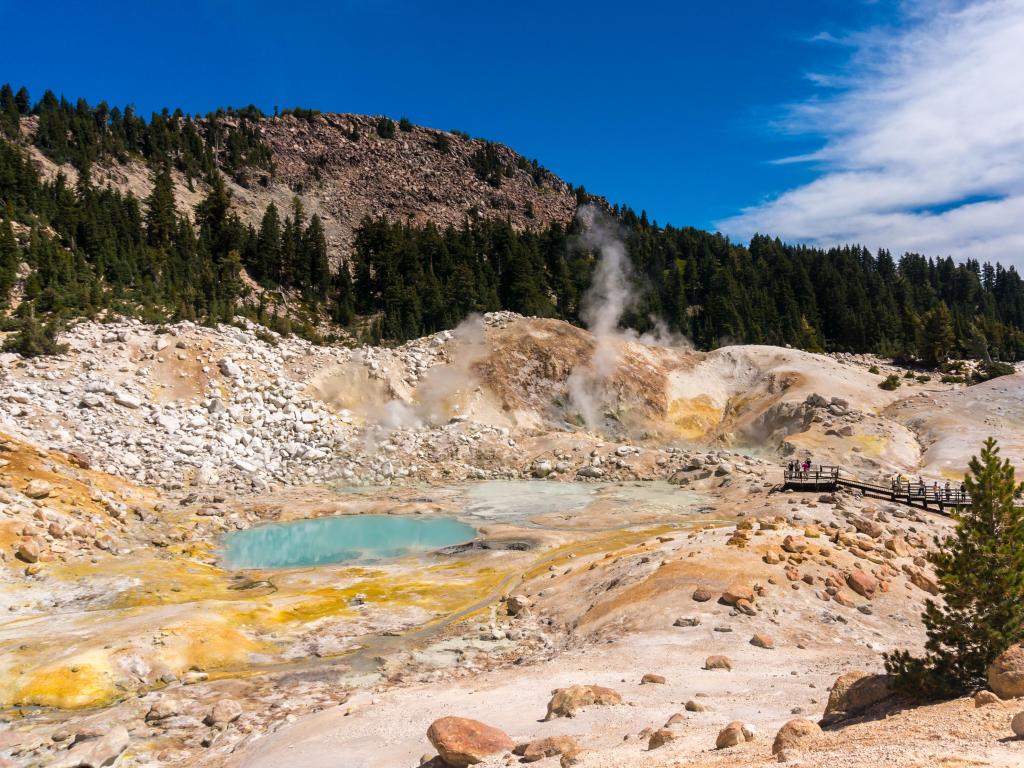 Lassen Volcanic National Park, California, USA taken at Bumpass Hell with volcanic terrain and a small water hole, rocky mountains and trees in the distance on a sunny day.