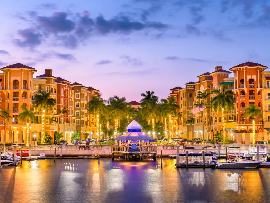 Skyline of downtown Naples, Florida at dusk. Small boats are moored on the quay in front of brightly lit palm trees.