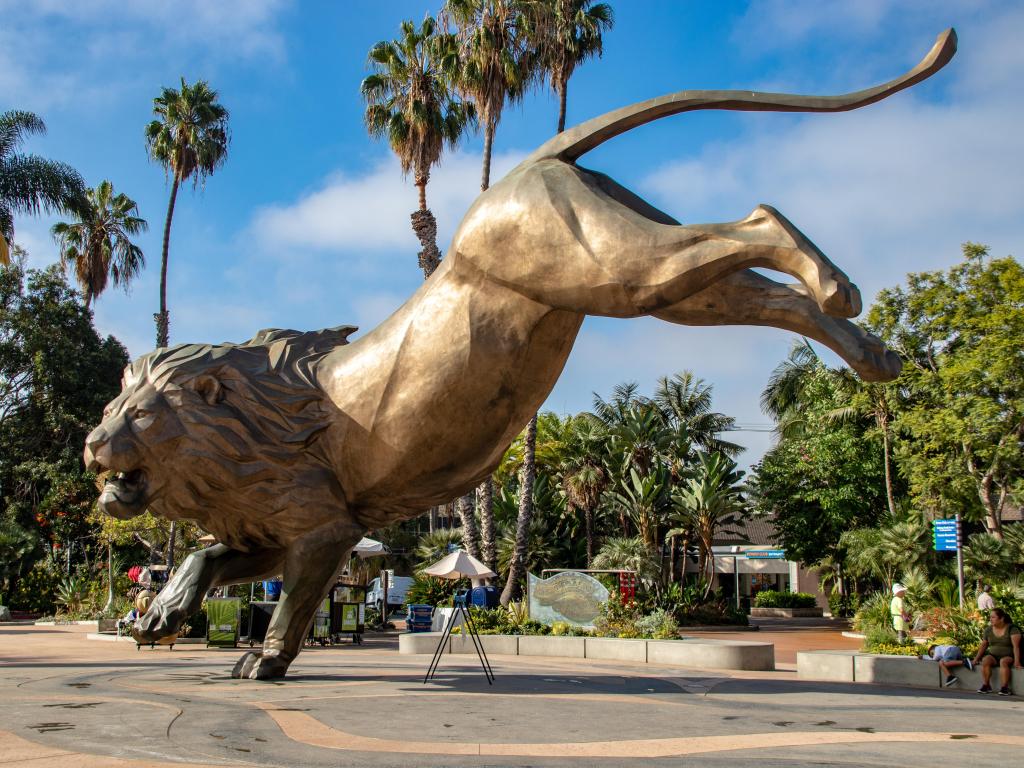 Lion statue at the entrance to San Diego Zoo with palm trees in the background and a blue sky.