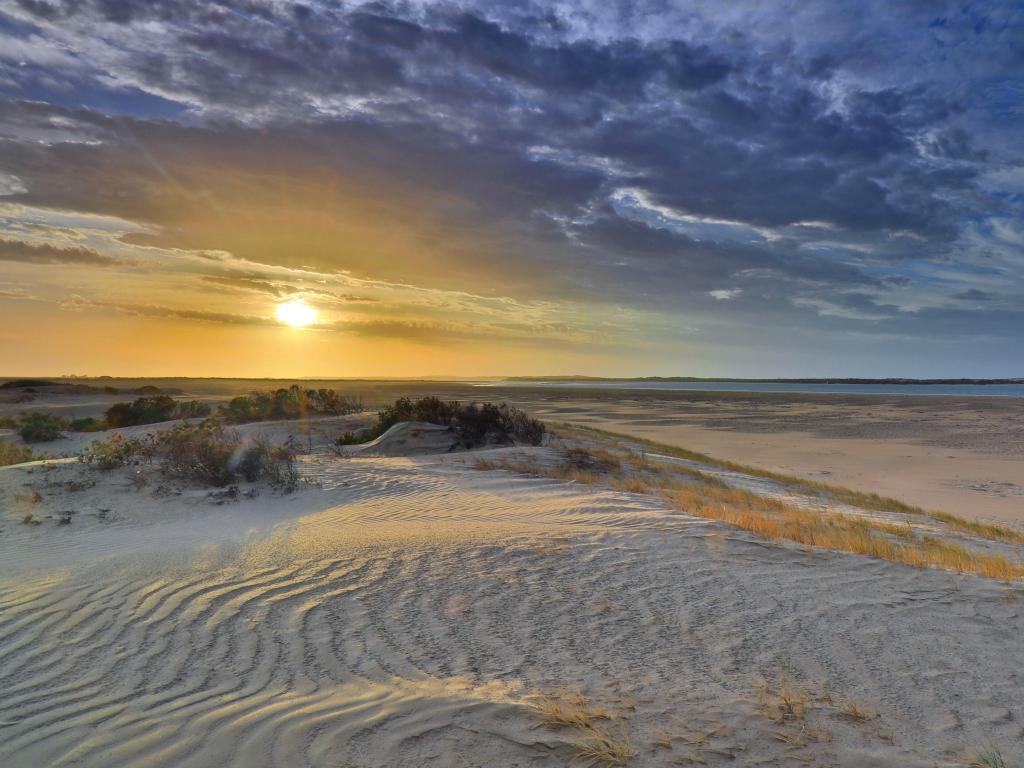 Flat, sandy landscape with calm ocean behind and setting sun