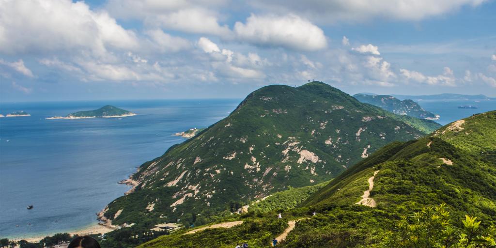 People walking up a hill on the Dragon's Back hiking trail, with a turquoise sea in the background 