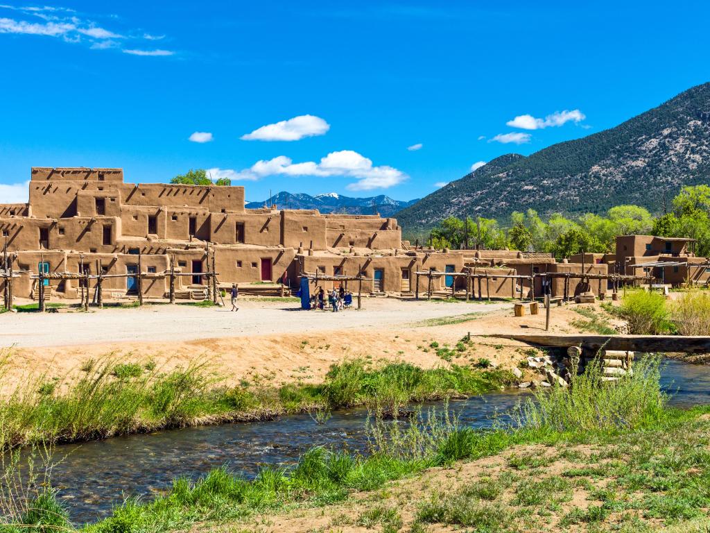 Taos Pueblo, New Mexico, USA with a river in the foreground and mountains in the distance on a sunny day.