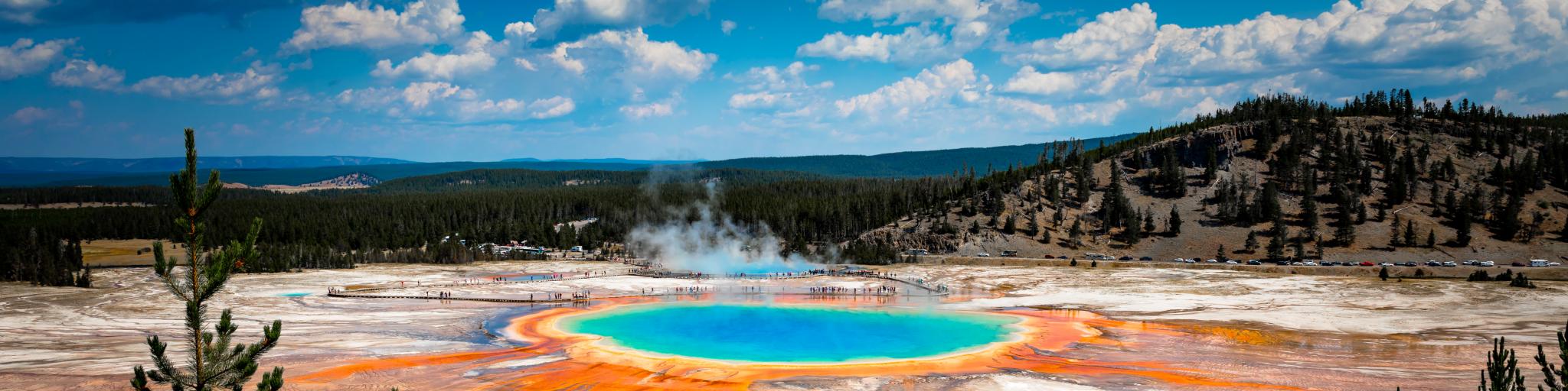 A panoramic view of the Grand Prismatic Spring in Yellowstone National Park show's that the water's color is a mix of orange, yellow, green, and blue.