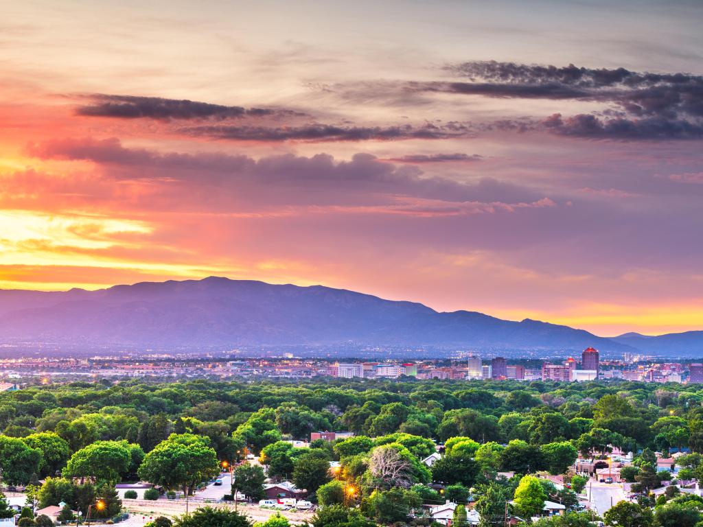 Albuquerque, New Mexico, USA with the downtown cityscape at twilight, green trees in the foreground and mountains in the distance.