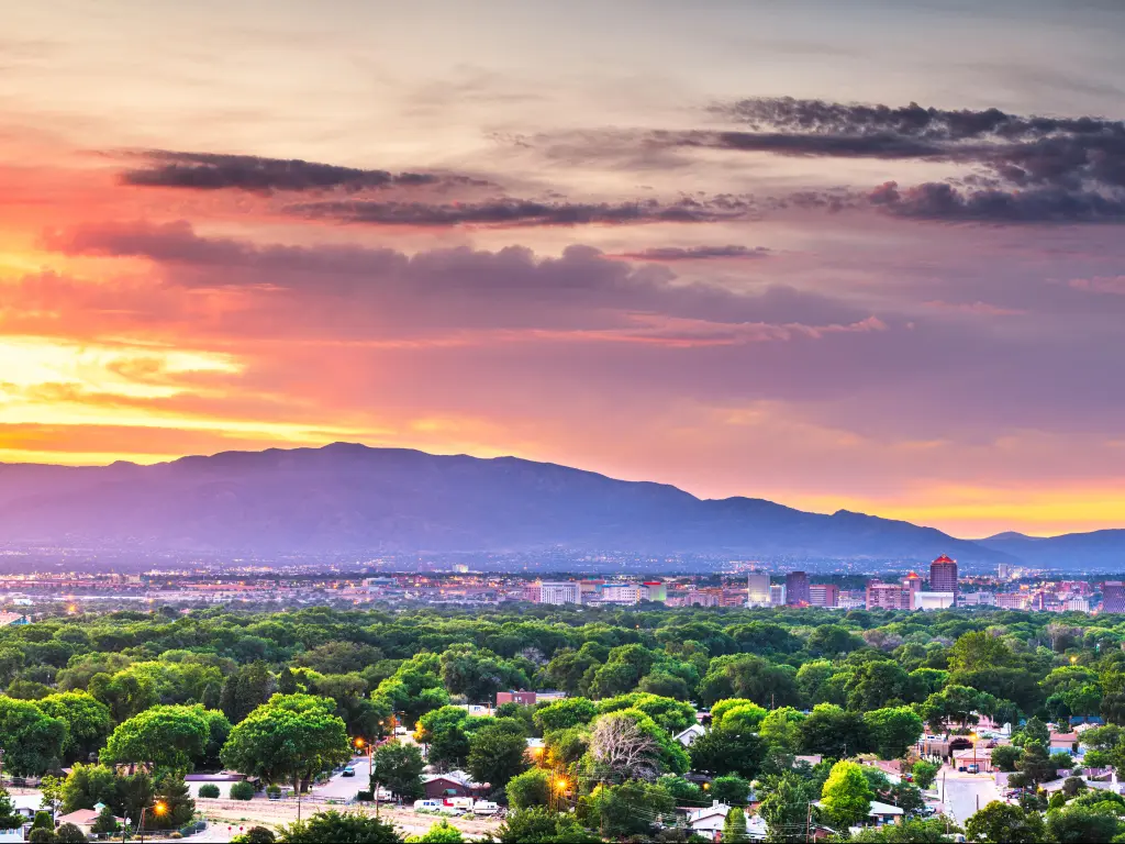 Albuquerque, New Mexico, USA with the downtown cityscape at twilight, green trees in the foreground and mountains in the distance.