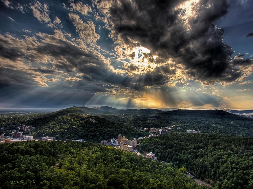 Hot Springs, Arkansas with a dramatic sky and sun rays through clouds above the city surrounded by large tree covered hills in the foreground and background.