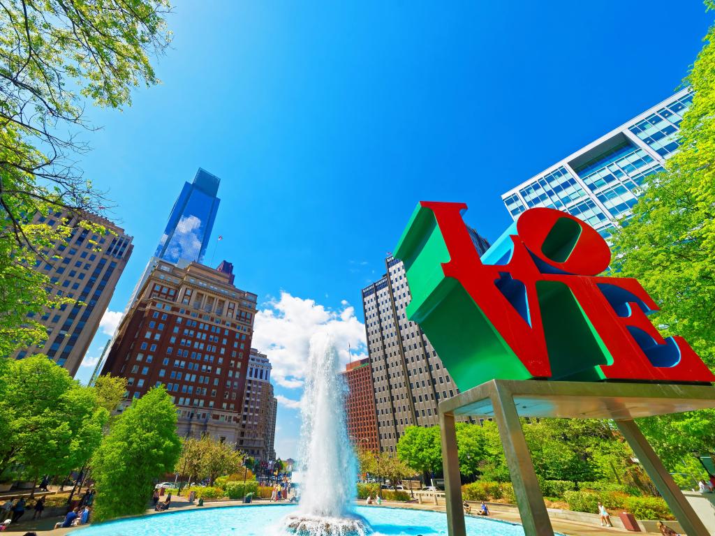  Love sculpture in the Love Park in Philadelphia, Pennsylvania, USA. Tourists in the park. Skyline with skyscrapers on the background