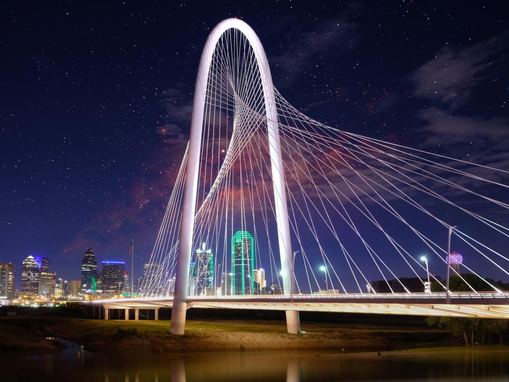 White metal bridge lit up in the foreground with high rise down town buildings behind and stars