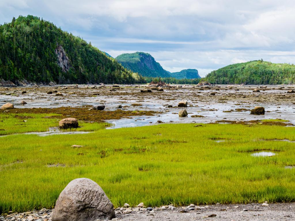 Bic National Park, Quebec, Canada with a grass foreground, water and rocks and then green mountains in the distance on a sunny but cloudy day.