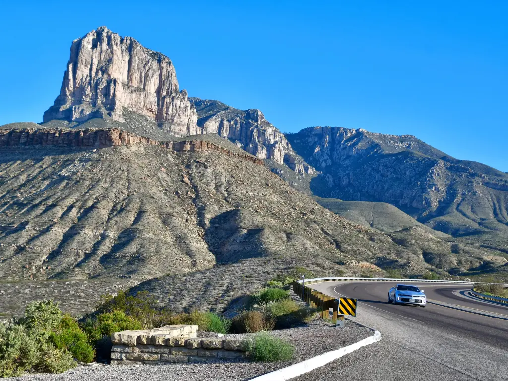 Rocky peak rises up above road with one car, with clear blue sky behind