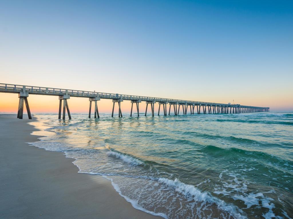 Panama City Beach, Florida with the a pier leading in the distance at sunrise with the beach and calm water in the foreground.