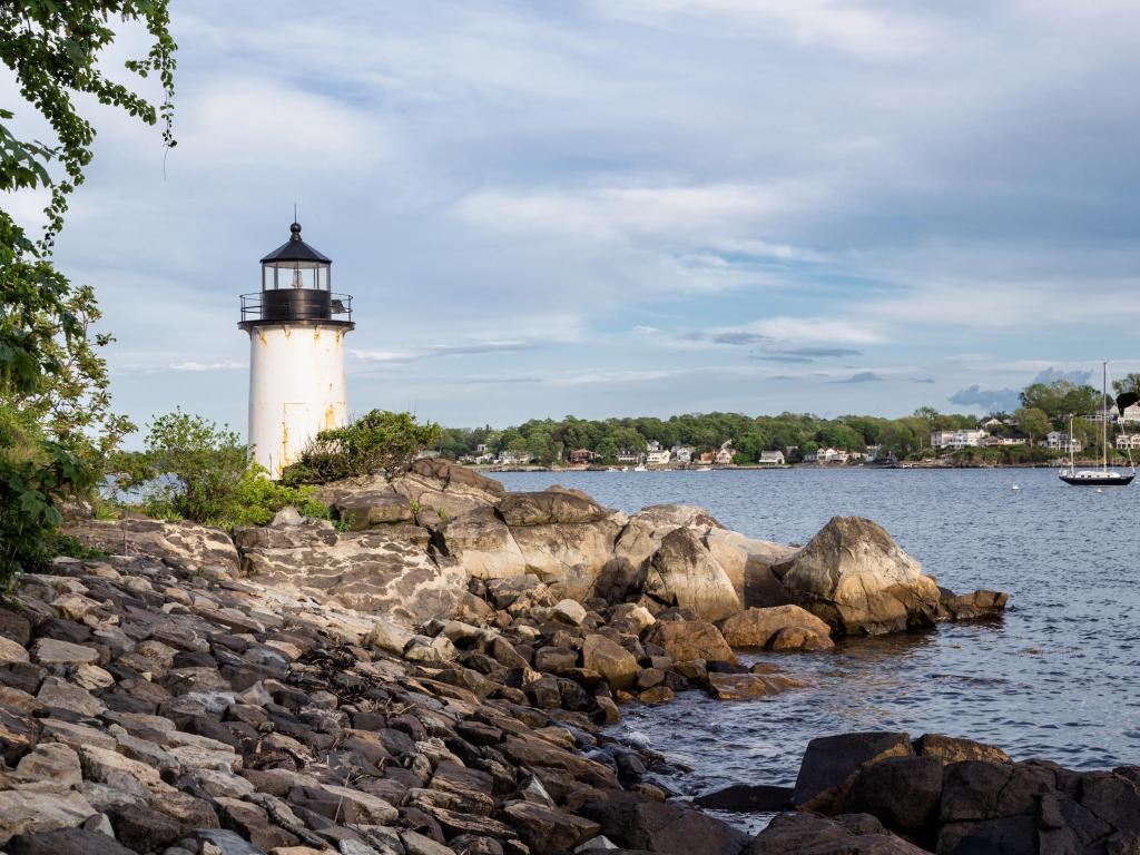 Salem, Massachusetts, USA with Winter Island Lighthouse behind rocks in the foreground, the sea surrounding and trees and buildings in the distance.