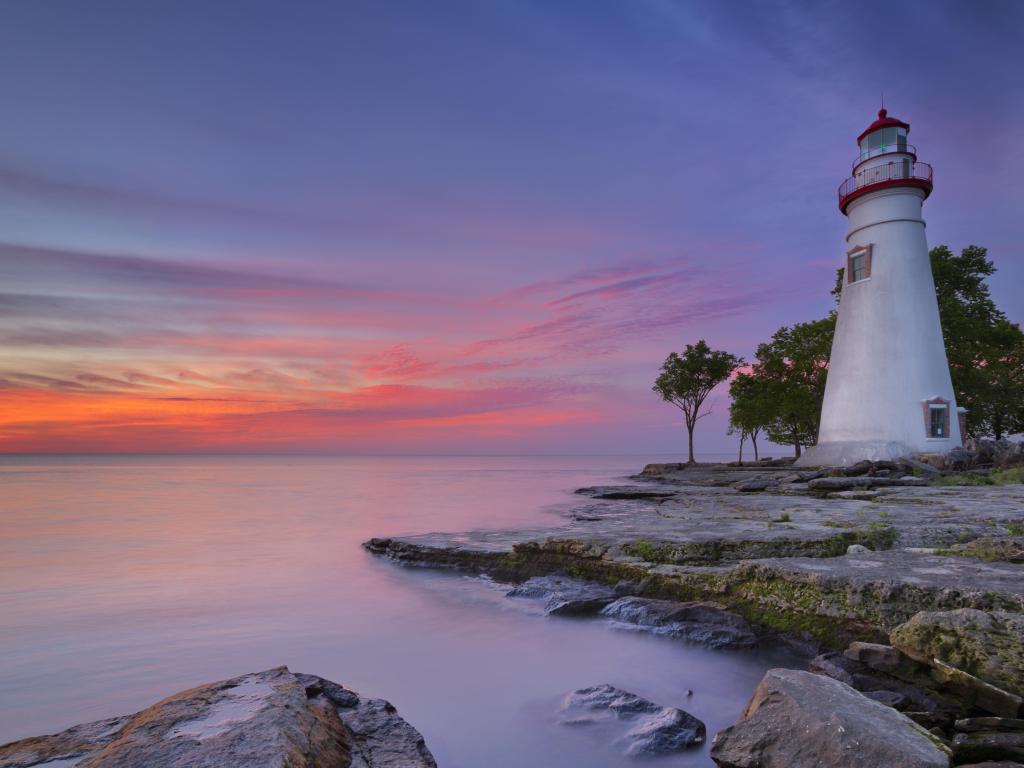 White lighthouse on a shoreline of flat rocks with calm, silver-grey lake water and pink sunrise