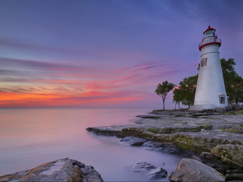 White lighthouse on a shoreline of flat rocks with calm, silver-grey lake water and pink sunrise