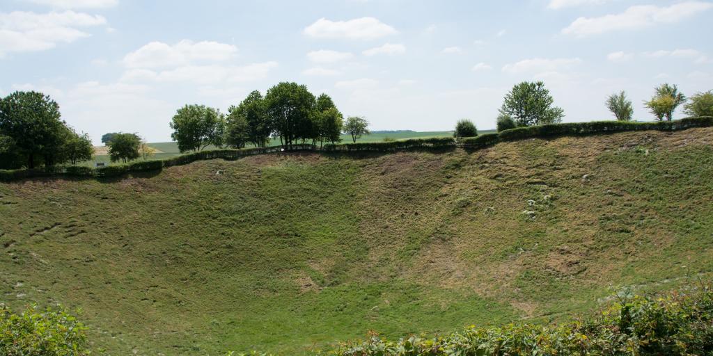 A section of the Lochnagar Crater, France, now covered in grass, with a hedge in front and trees behind